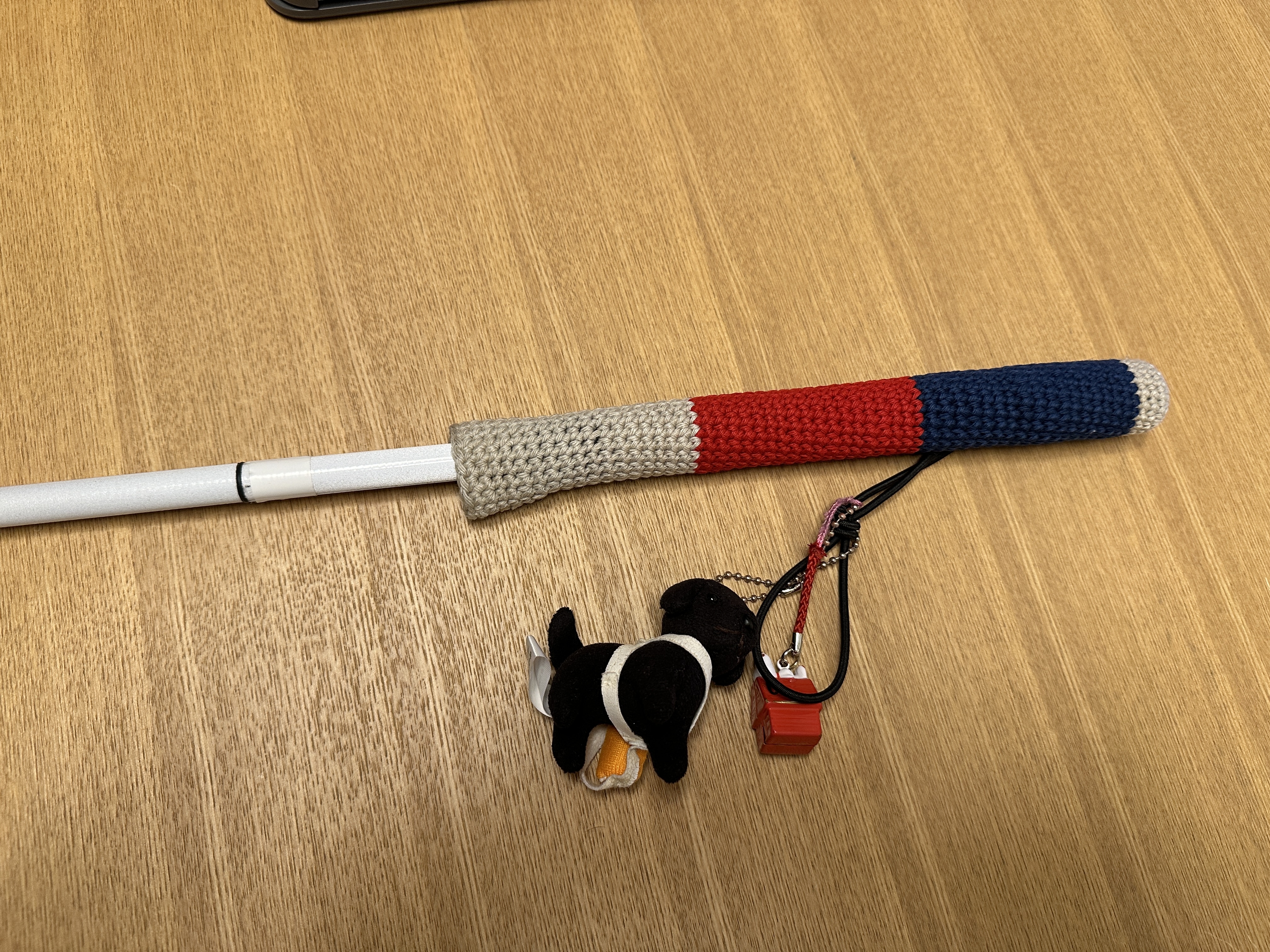 トリコロールのニットカバーが被せられた、白杖の持ち手の写真。ストラップ部分には盲導犬のマスコットとスヌーピーの鈴が付けられています。