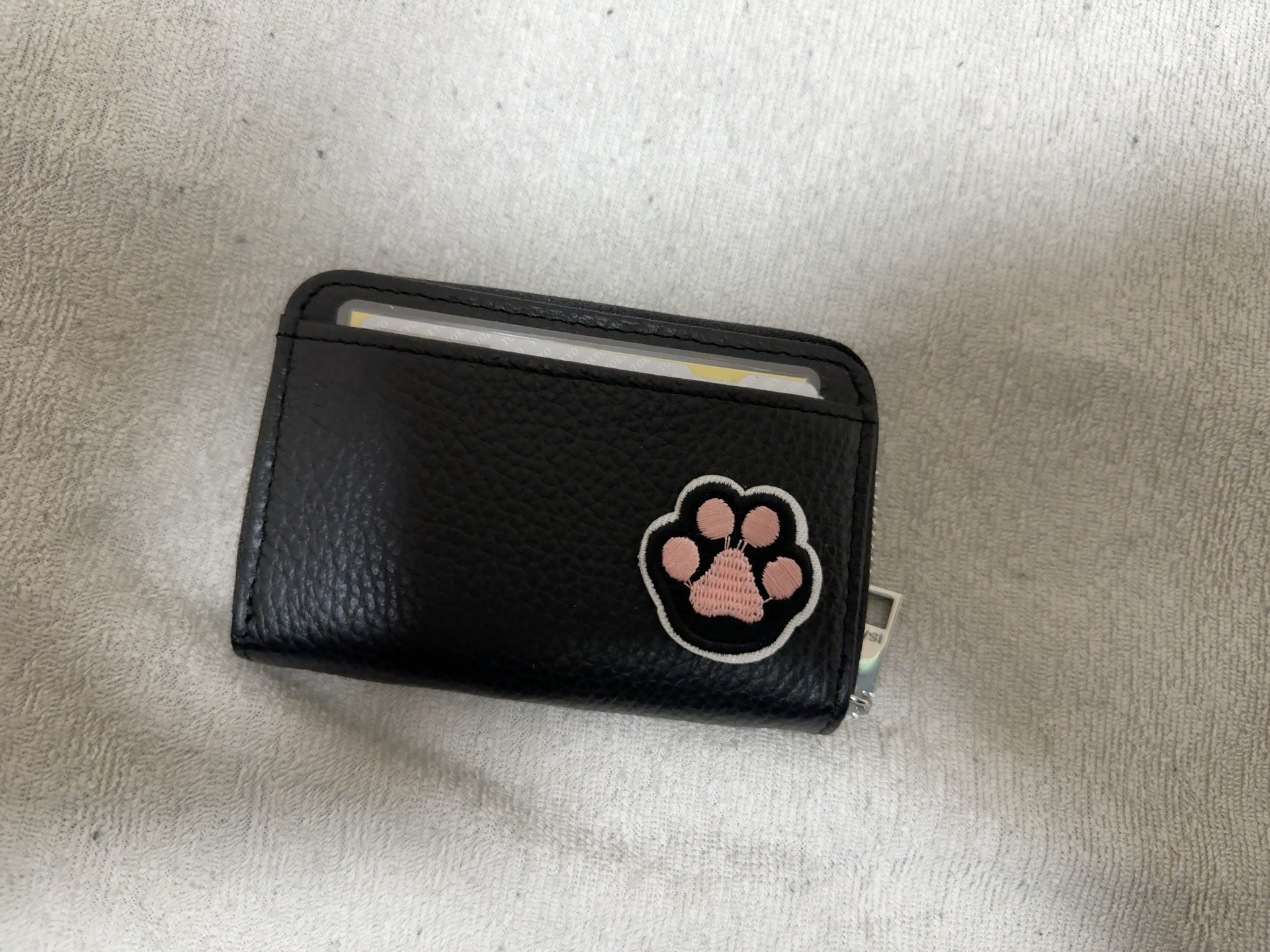 黒い革製のカードケースを写した写真。表面にはシボ加工が施されており、更に黒猫の手にピンク色の肉球が詩集されたアップリケが貼り付けられています。